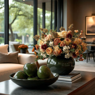 Bowl of small green glazed stoneware apples styled on coffee table with large floral arrangement in deep green vase in modern living room setting.
