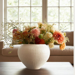Handcrafted white crosshatch ceramic pot filled with vibrant faux dahlias, ranunculus, and greenery on a wood coffee table in a sunlit living room