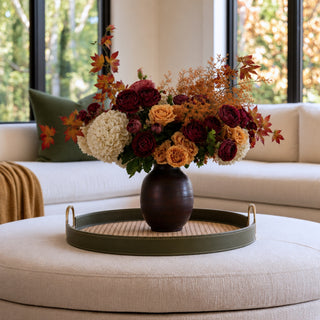 Warm modern transitional living room with a cream sectional, olive green linen pillows, honey brown throw blanket, and fall floral arrangement styled in a dark wood vase on a 20-inch olive green leather and rattan tray. Cozy autumn home decor with burgundy, rust, orange, cream, and amber tones.