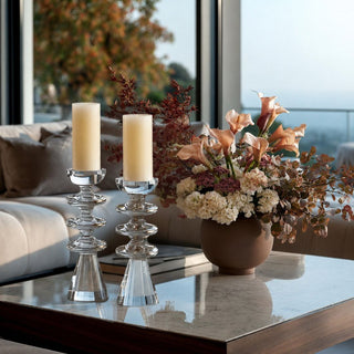 Two clear glass candlestick holders with cream pillar candles on a marble coffee table in a modern living room, next to a terracotta vase filled with peach and cream flowers.