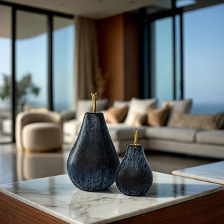 Two dark blue frosted glass pears with gold stems, displayed on a white marble-topped table in a modern, sunlit living room.