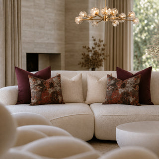 A modern cream bouclé sectional sofa in a luxury living room setting, styled with deep burgundy and textured rust-patterned throw pillows. A gold multi-globe chandelier hangs above, and a blurred foreground chair adds depth.
