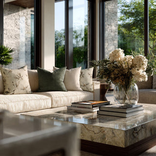 A modern living room scene with a cream ribbed sofa, willow foliage green and print design decorative throw pillows, a marble coffee table, and a glass vase of white flowers in front of large windows.