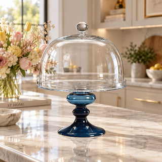 The Bellissima Glass Cake Stand featuring a clear glass serving plate and domed cover with a deep blue glass pedestal base, styled on a marble kitchen island in a bright luxury kitchen.