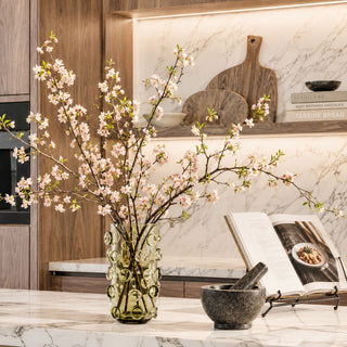 11.75-inch olive green decorative glass vase featuring sculptural hand-blown bubble accents, styled with flowering cherry blossom branches on a white marble countertop. The transparent olive glass reveals the stems inside, creating a sophisticated luxury floral arrangement in a contemporary designer kitchen.