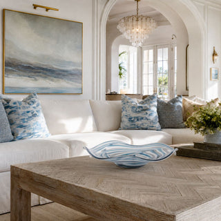 15.75-inch Blue Ocean Swirl glass bowl centerpiece on a rustic oak coffee table in a luxury coastal living room. This handcrafted blue and white art glass bowl features a wavy organic silhouette and ocean-inspired swirl pattern, styled within an elegant California coastal home with designer furnishings and refined neutral décor.