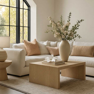 Matte white porcelain hibiscus flower styled on a stack of books atop a natural wood coffee table in a modern living room with cream-colored sofa and soft peach accents