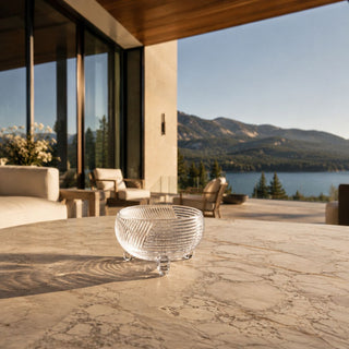 Clear ribbed glass footed bowl styled on a limestone outdoor coffee table at a luxury Lake Tahoe patio, illuminated by warm summer sunlight and overlooking contemporary outdoor living spaces.