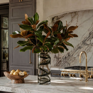Third & Main 13-inch smoked olive ribbon glass vase styled with magnolia branches on a marble kitchen island in an elegant luxury kitchen featuring brass accents and soft charcoal cabinetry.
