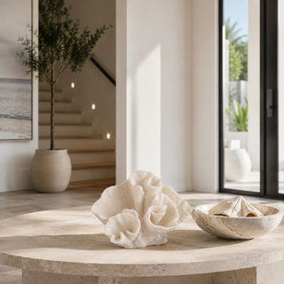 White sculptural coral décor displayed on a clear acrylic stand atop a round travertine coffee table in a luxury coastal-inspired home. Warm natural sunlight, an olive tree, floating wood staircase, and neutral Mediterranean-style interiors create an elegant, serene backdrop that highlights the organic texture and museum-quality design of the coral centerpiece.