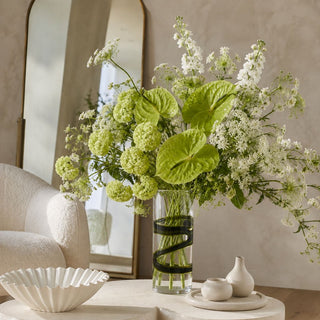 Green and white luxury floral arrangement in a clear smoked ribbon glass vase with visible stems, styled on a sculptural ivory coffee table in a contemporary neutral living room.