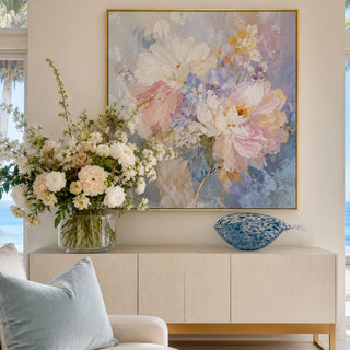 Third & Main blue seaside glass fish bowl displayed on a luxury coastal console table beneath a pastel floral artwork, featuring handblown blue speckled art glass, organic fish-inspired shape, and elegant beach house styling in a bright oceanfront living room.