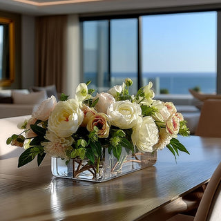 True Bloom 25 Inch Cafe Au Lait Real Touch premade floral arrangement styled on a modern dining table in a luxury Malibu coastal home with floor-to-ceiling windows and ocean views, featuring lifelike ivory peonies, roses, hellebores, and blush accents arranged in a thick-cut rectangular glass vase with permanent botanical resin.