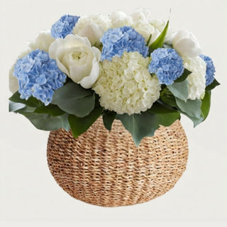 True Bloom Blue Hydrangea Garden Arrangement displayed in round woven seagrass vase featuring blue and white real touch hydrangea and white roses