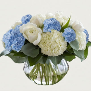 True Bloom Blue Hydrangea Garden Arrangement in clear sculpted glass bowl with visible stems and floral pebbles, featuring layered blue hydrangea and white roses on neutral background