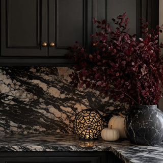 Black spiderweb water-spinning LED globe glowing with silver sparkle beside cream pumpkins and dark marble decor in a Halloween kitchen vignette.