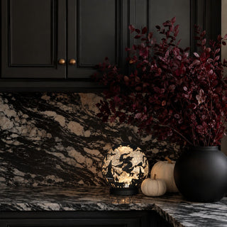 Black witch water-spinning LED globe glowing with silver sparkle beside cream pumpkins, burgundy foliage, and black marble kitchen decor.