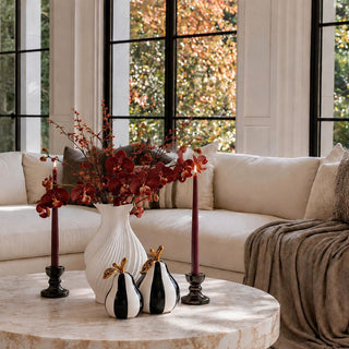 Black and white striped ceramic pears styled on a travertine coffee table with a white fluted vase, burgundy orchids, and plum taper candles.