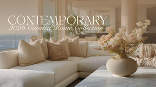 Bright contemporary living room featuring a neutral sectional sofa layered with textured beige pillows, a sculptural stone vase filled with soft blush and ivory florals, and a minimalist modern interior with natural light and ocean views