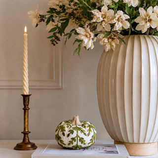 Small cream and olive foliage knit pumpkin styled on books beside a brass taper candleholder and cream floral vase in a warm neutral interior.