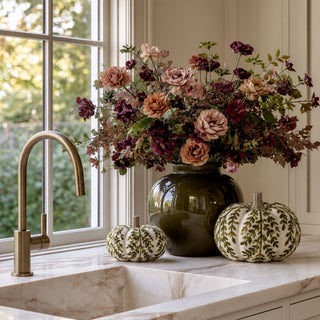 Medium and large cream and olive foliage knit pumpkins styled beside a dark green vase and fall floral arrangement on a marble kitchen counter.