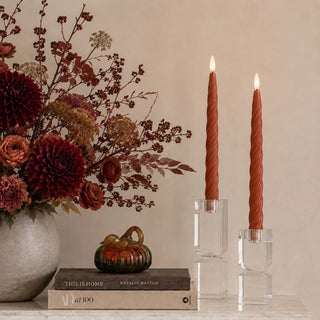 Styled set of two 9.75 inch Ember Spiral LED taper candles in rust-orange wax with spiral texture and flameless warm light, displayed in clear holders beside burgundy and rust fall florals on a marble tabletop.