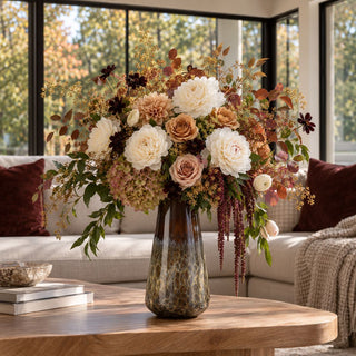Large 14-inch brown and cream blown glass vase filled with ivory peonies, blush and caramel roses, burgundy flowers, and autumn foliage on a wooden coffee table in a contemporary living room.