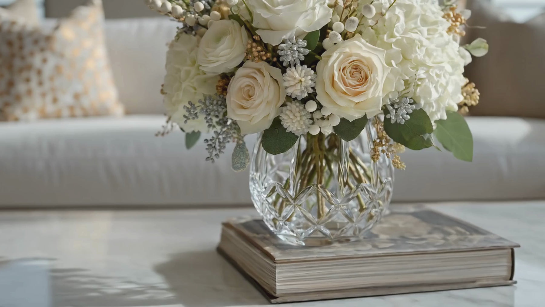 A beautiful True Bloom Real Touch Faux Floral Arrangement of Champagne Duchess Roses and other complementary florals in a cut glass vase on the coffee table of a luxury decor style living room 