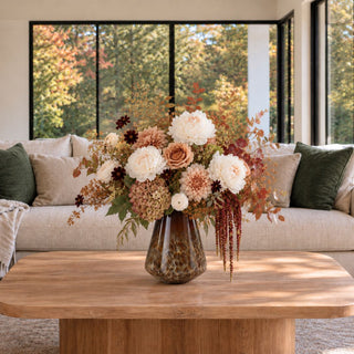 Small 11-inch brown and cream blown glass vase styled with ivory flowers, muted blush roses, burgundy accents, and autumn branches on a wood coffee table in a warm neutral living room.