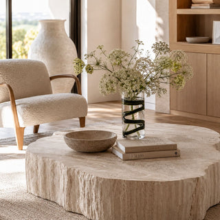 Third & Main 9 Inch Smoked Olive Ribbon Glass Vase displayed on a sculptural faux-limestone coffee table in a sunlit luxury coastal living room. The clear cylindrical glass vase features bold smoked olive ribbon-like swirls wrapped around the vessel, allowing the flower stems to remain visible through the transparent glass.