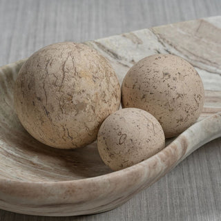Three natural travertine stone decorative orbs in graduated sizes styled in a warm gold bowl on a marble tabletop with crystal taper candle holders.